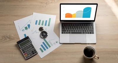 Desk flat lay with a laptop showing a revenue growth chart, printed forecast charts, a compass, and a calculator, representing sales forecasting methods